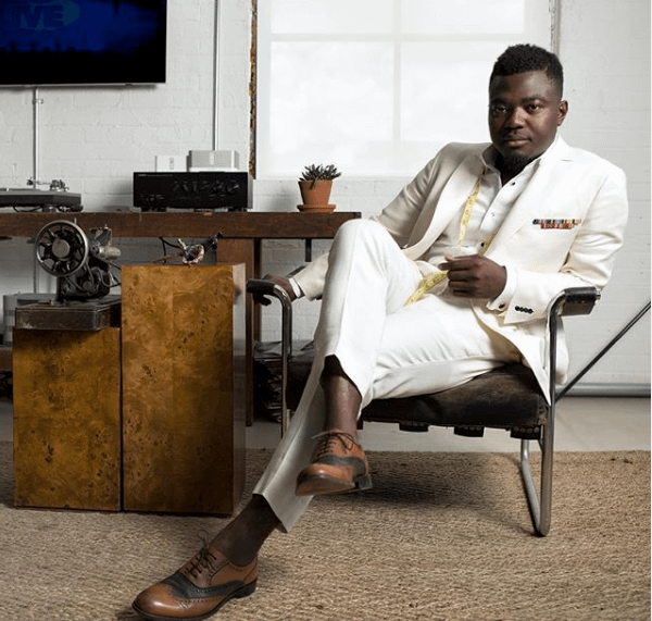 pictured: nana Bediako seated in a white suite with a sewing machine on a desk beside him